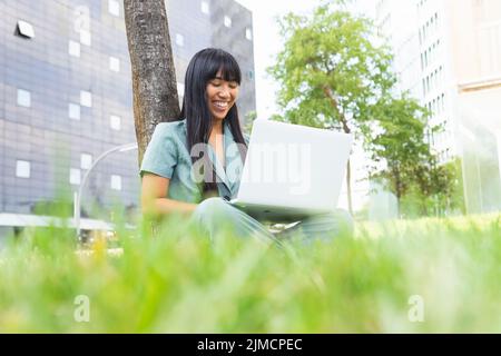 Young smiling woman with long hair sitting on grass near tree and using netbook while doing homework on summer day in park Stock Photo