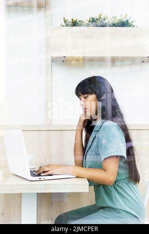 Young Asian woman with long hair typing on laptop keyboard while sitting at table behind glass and working on remote project in cafeteria Stock Photo