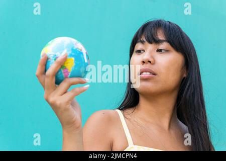 Thoughtful young ethnic female tourist with long hair examining small globe while planning trip against turquoise background Stock Photo