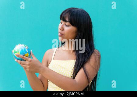 Thoughtful young ethnic female tourist with long hair examining small globe while planning trip against turquoise background Stock Photo