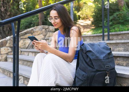 Positive female student texting message on cellphone while sitting on staircase with backpack on street of city on summer day Stock Photo