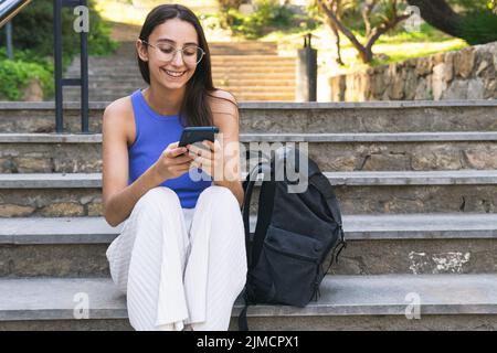 Positive female student texting message on cellphone while sitting on staircase with backpack on street of city on summer day Stock Photo
