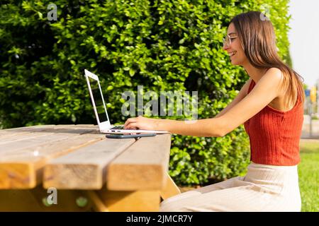 Side view of female student typing on netbook while doing homework in park with trees on summer day Stock Photo