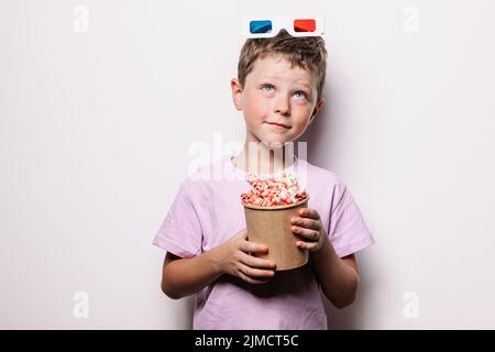 Boy with modern 3D glasses holding cardboard bucket with popcorn while looking up against white background in studio Stock Photo