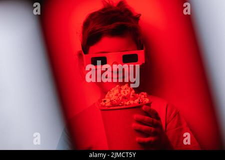 Through red lens of content girl in 3D glasses with tasty popcorn standing against gray background in light studio