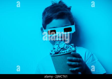 Through red lens of content girl in 3D glasses with tasty popcorn standing against gray background in light studio