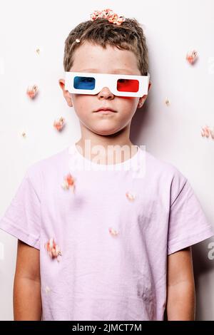 Cute kid in 3D glasses with colorful lenses throwing popcorn over head while looking at camera standing against white background in light studio