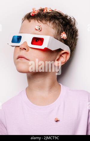 Cute kid in 3D glasses with colorful lenses throwing popcorn over head while looking away standing against white background in light studio