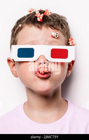 Cute kid in 3D glasses with colorful lenses eating tasty popcorn while standing against white background in light studio