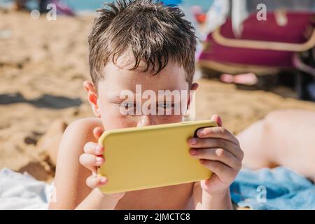 Focused boy in swimming shorts playing video game on cellphone while lying on sandy shore in tropical resort on sunny summer day in Getaria Zarautz