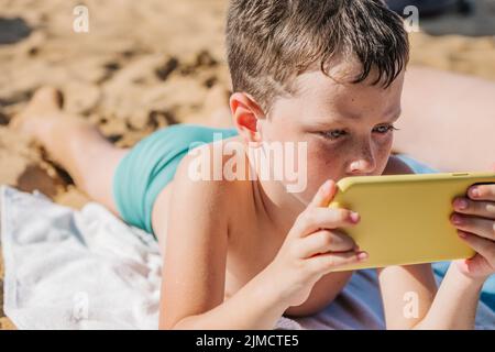 Focused boy in swimming shorts playing video game on cellphone while lying on sandy shore in tropical resort on sunny summer day in Getaria Zarautz