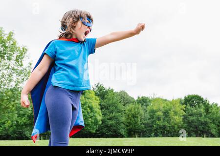 Side view of little female superhero with cape and mask raising arm with clenched fist and screaming against green trees and cloudy sky on summer day Stock Photo