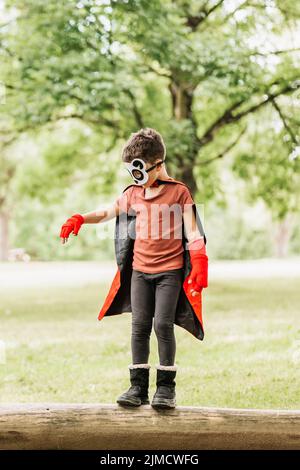 Full body cute little superhero boy with panda mask and cape walking on log while playing in green park in summer