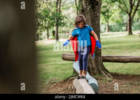 Full body cute little superhero with cape and gloves walking on log while playing in green park in summer