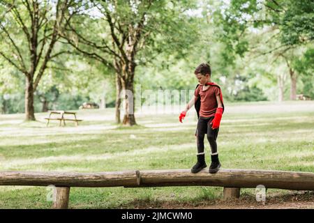 Full body cute little superhero with cape and gloves walking on log while playing in green park in summer Full body cute little superhero with cape and gloves walking on log while playing in green park in summer