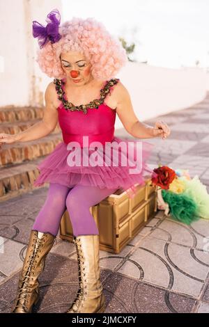 Concentrated middle aged female circus artist in funny costume and pink wig with bright makeup and fake nose sitting on chair before performance on st Stock Photo