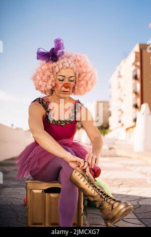 Concentrated middle aged female circus artist in funny costume and pink wig with bright makeup and fake nose sitting on chair and putting on boots bef Stock Photo