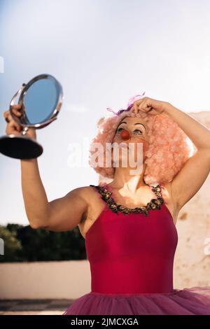 Low angle of confident mature female clown in bright costume and funny curly wig with makeup looking in round cosmetic mirror while standing on street Stock Photo