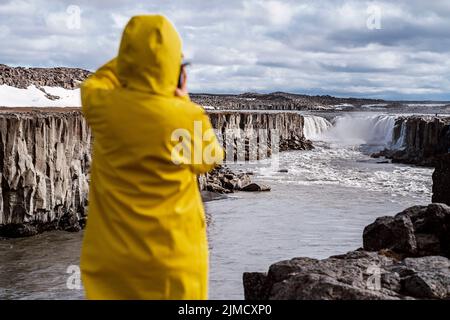 Back view of unrecognizable traveler in bright yellow hooded raincoat taking photo of amazing scenery of Selfoss waterfall and river flowing in gorge Stock Photo