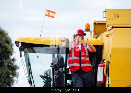 Positive female combine operator in uniform showing thumb up gesture and looking at camera while standing near industrial harvester in countryside Stock Photo