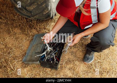 From above of crop unrecognizable female worker in uniform choosing tools from box while hunkering down on agricultural field in countryside Stock Photo