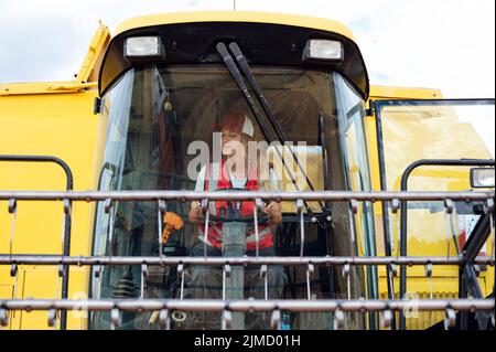 Focused female combine operator in uniform harvesting grains with professional machine in agricultural plantation while working in countryside on summ Stock Photo