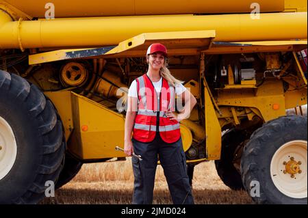Cheerful female worker in uniform with wrench looking at camera with smile while standing near combine harvester in agricultural field Stock Photo