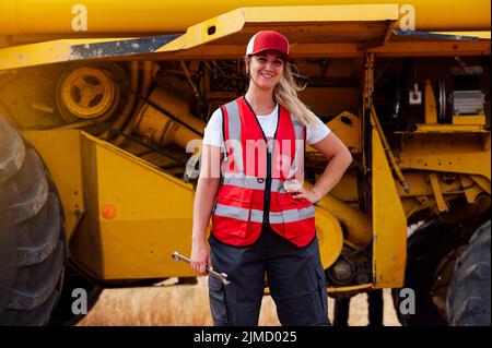 Cheerful female worker in uniform with wrench looking at camera with smile while standing near combine harvester in agricultural field Stock Photo