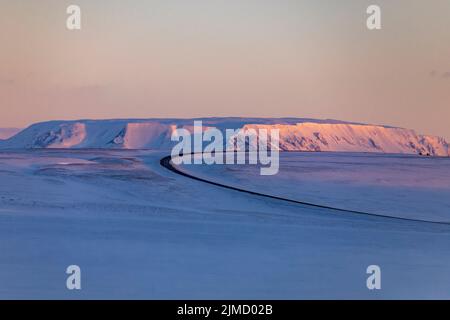 Empty winding asphalt roadway leading through valley covered with snow in highlands during sunset in winter in Iceland Stock Photo