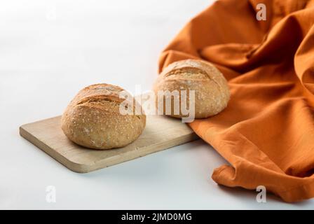 Fresh loaves of bread served on wooden chopping board near orange fabric on white background Stock Photo