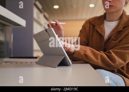 Unrecognizable woman with stylus making notes on tablet while doing homework assignment in library in daytime Stock Photo