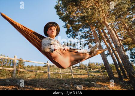 Pensive male in hat lying in hammock while resting in forest with green trees on sunny summer day Stock Photo