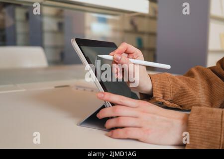 Unrecognizable woman with stylus making notes on tablet while doing homework assignment in library in daytime Stock Photo