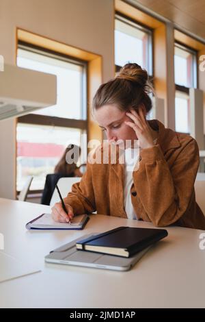 Young woman in casual clothes with hair bun touching head and writing in planner while sitting at table and studying in university library Stock Photo
