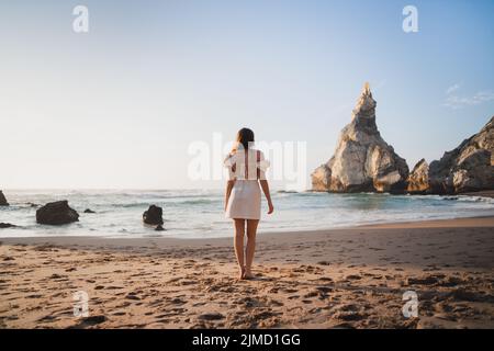 Back view of young female traveler in dress standing on sandy shore with rough rocky cliffs near sea on summer vacation Stock Photo