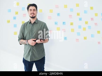 I have big plans for my career. Portrait of a young businessman using a digital tablet while standing against a wall with notes in an office. Stock Photo