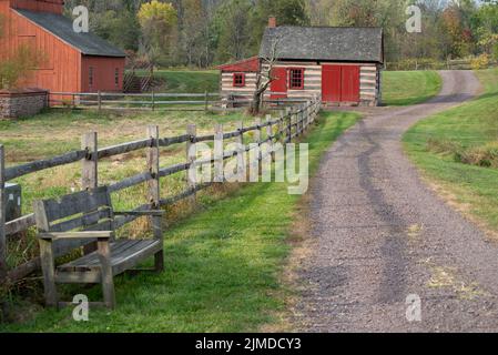 Gravel path leads past an empty bench, red barn and log cabin Stock Photo