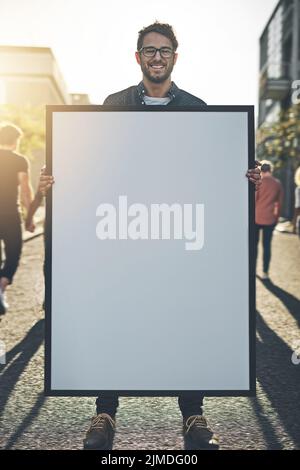 Young man holding an empty sign, board or placard while standing outside in the city street. Portrait of one happy, smiling and cheerful guy making an Stock Photo