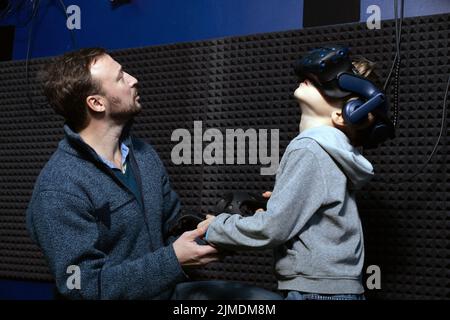 Man helps boy in virtual reality helmet with joysticks in his hands understand how to control video game Stock Photo