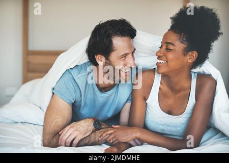 Happy, carefree and laughing couple having fun lying in bed together. Interracial husband and wife bonding and showing affection while talking Stock Photo