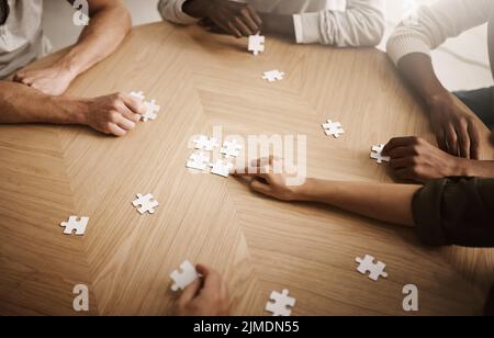 Friends hands solving a puzzle in a group collaboration together as a team on wooden table indoors. Diverse colleagues using teamwork and support to Stock Photo