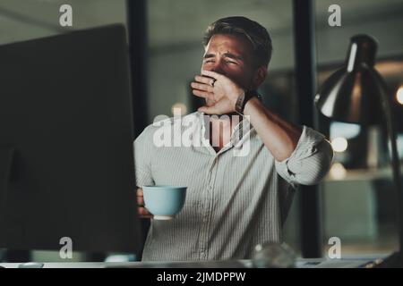 Maybe coffee will keep me awake. a young businessman yawning while working on a computer in an office at night. Stock Photo