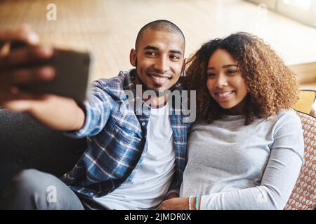 Smile babe. Happy, romantic and in love couple taking a selfie together on phone sitting on couch. Smiling young spouses hugging while holding phone Stock Photo