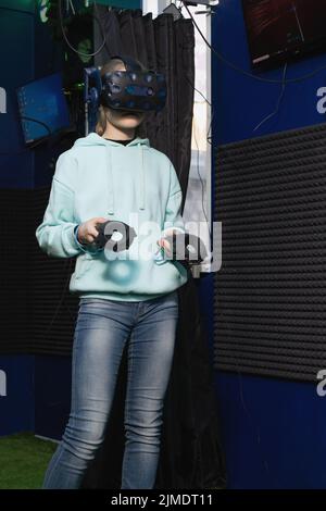 Teenage girl in virtual reality helmet tries to control game using controllers in VR club Stock Photo