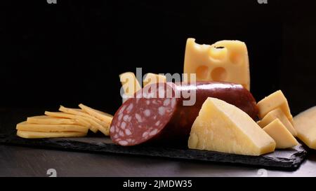 Several types of cheese and piece of salami loaf are on black stone board. Stock Photo