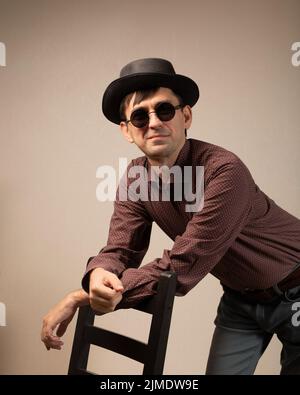 Smiling flirtatious man in black hat and black glasses leaning his arm on back of chair. Stock Photo