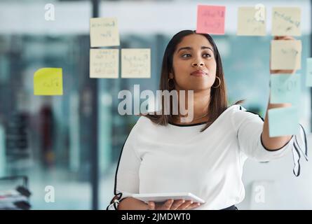 Building the best plans. a young businesswoman using a digital tablet while brainstorming with notes on a glass wall in an office. Stock Photo