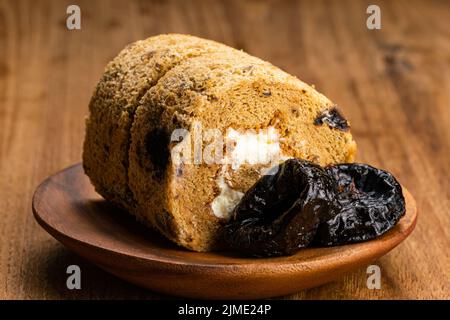 Closeup view of homemade prune sponge cake roll and dried pitted prune fruits in wooden plate. Stock Photo