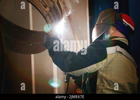 This is a welder in a protective mask and work clothes at work in the production shop. A genuine working environment. Stock Photo