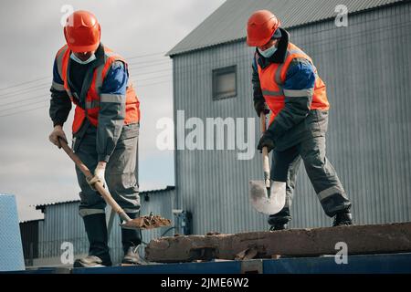 Two workers in hard hats, work clothes and a medical mask work with shovels at a construction site. Hard physical labor. Stock Photo